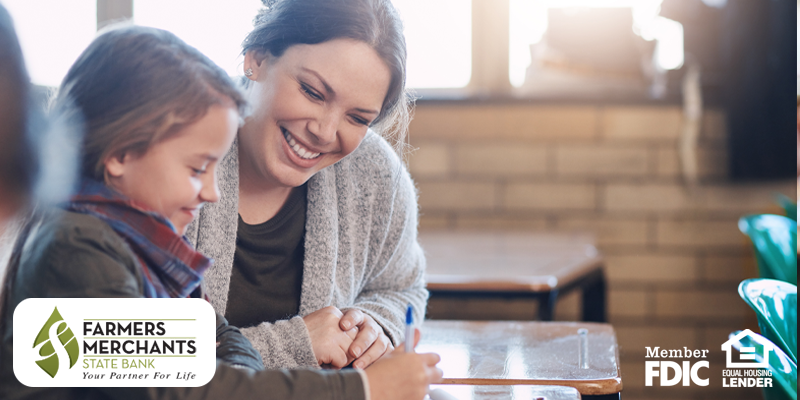 Image shows girl writing at desk with smiling teacher beside her
