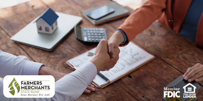 Lender and homebuyer shaking hands across table with clipboard, laptop, calculator and house figures present