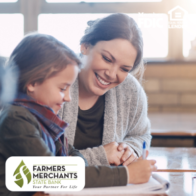 Image shows girl writing at desk with smiling teacher beside her