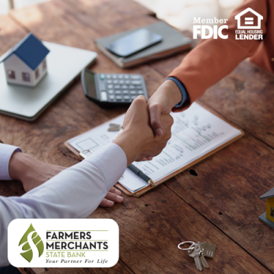 Lender and homebuyer shaking hands across table with clipboard, laptop, calculator and house figures present