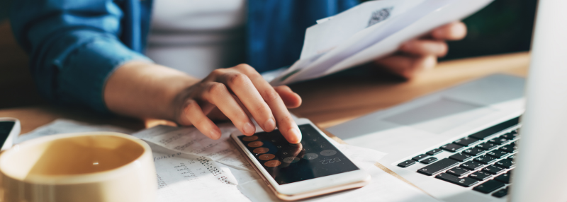Woman typing on smartphone calculator while holding papers in hand with laptop and mug on desk