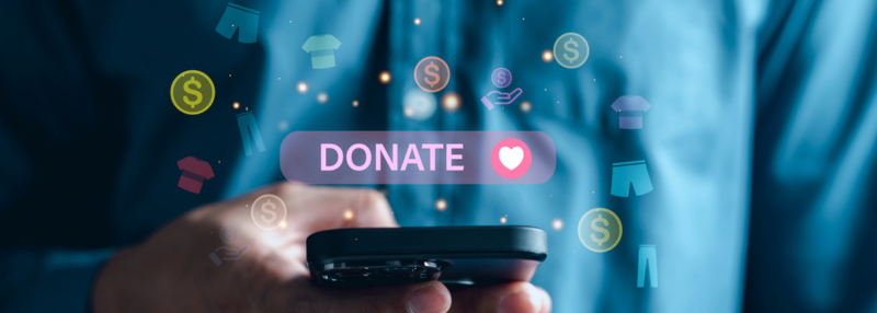 Hand holding smartphone with Donate text and heart icon hovering above it and money symbol and clothing icons floating around it