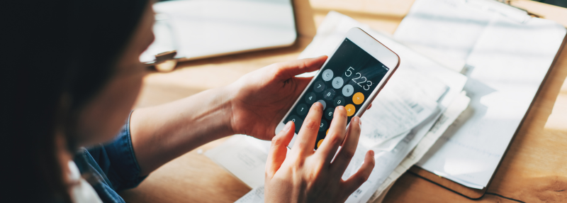 Woman using smartphone calculator with laptop and papers on the table behind