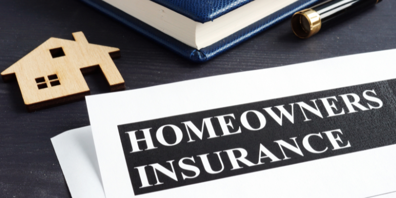 Paper with Homeowners Insurance written on it in caps with a wooden house block, journal and fountain pen in background