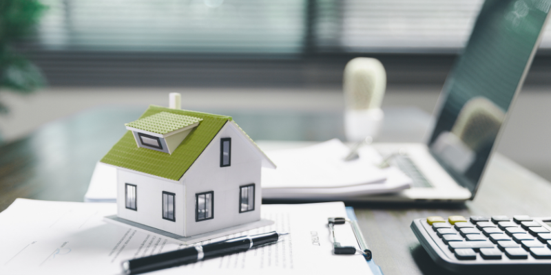 White house figurine with green roof on top of a clipboard with writing and a pen next to it, keyboard and laptop in background on desk blurred out