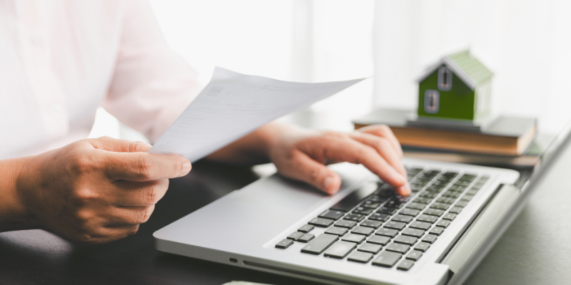 Man typing on laptop with paper in his hand and small house figurine on stack of books in background