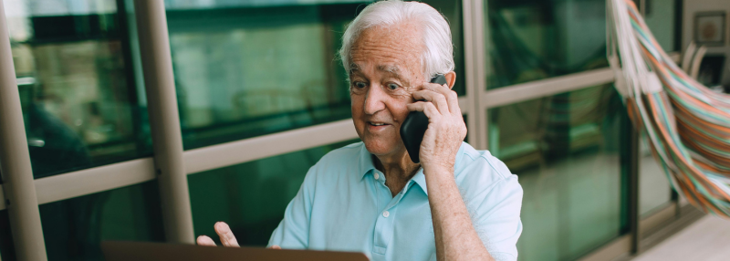 Senior man sitting in front of his laptop outside while talking on the phone